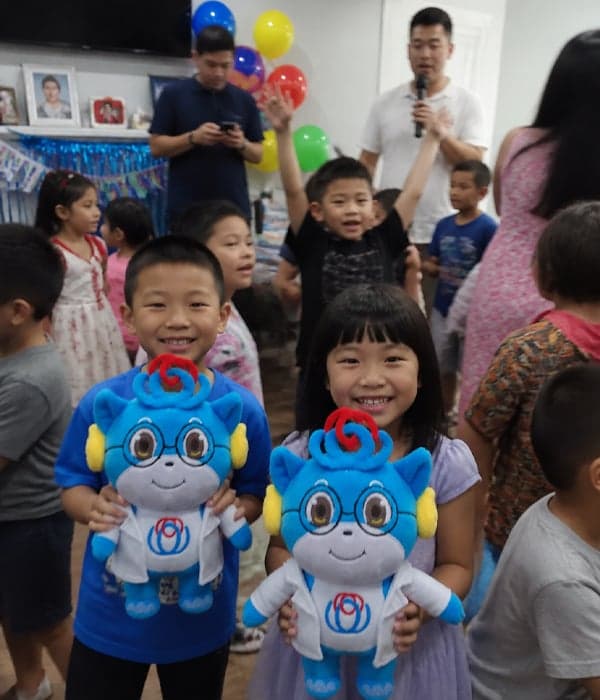Two smiling children holding matching custom blue cartoon character mascot plush toys with red curly horns and embroidered face at a kids' party event