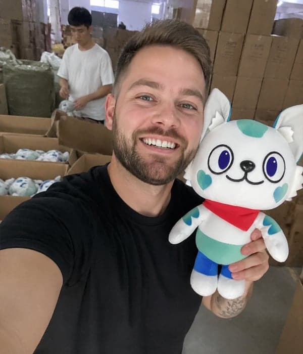 Client showcasing a custom white cat mascot plush with teal accents and red bandana on the StarDream Toys Shenzhen factory floor while staff pack the bulk order