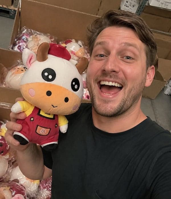 Buyer holding a custom cow character plush in red overalls and yellow shirt at the warehouse, with bulk-packed plush orders ready for shipment behind